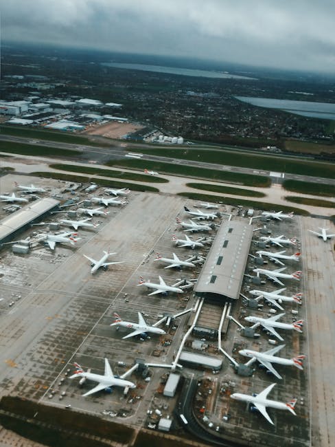 An aerial view of an airport tarmac showing numerous parked aircraft connected to jet bridges. The aircraft are mainly white with some featuring red markings on their tails. Surrounding the planes are ground support vehicles, cargo containers, and airport equipment. Beyond the runway, there are large open areas with some structures and a distant view of the cityscape under a cloudy sky. The image captures the busy scene typical of aircraft handling and terminal operations, relevant to home relocation and packing processes supported by Hounslow Man and Van, which offers removals services in the Heathrow area.