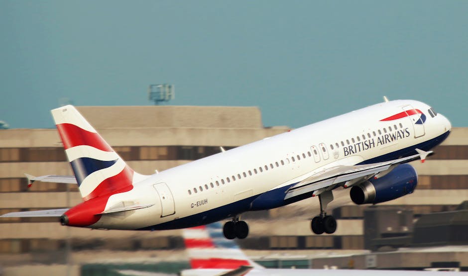 A British Airways commercial airplane is captured mid-takeoff on the runway, with its landing gear still extended and slightly blurred due to motion. The aircraft's white fuselage features the airline's name and logo, with red, white, and blue accents visible on the tail fin and engines. In the background, there is an industrial building with a flat roof, some lighting fixtures, and a clear sky above, indicating a typical airport setting. The airplane is in the process of ascending, and the image emphasizes aircraft movement related to home relocation logistics, connecting to furniture transport and packing and moving services by Hounslow Man and Van, especially in the Heathrow area near the airport in Hounslow.