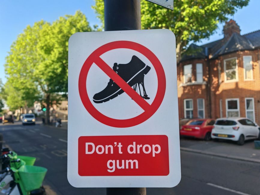 Close-up view of a rectangular traffic sign attached to a metal pole in a residential street. The sign features a black silhouette of a shoe with chewing gum stuck underneath, crossed out with a red circle and diagonal line. Below the symbol, there is a red rectangular box with white text that reads, 'Don't drop gum.' In the background, there are vehicles parked along the curb, including a white car and a red car, with a row of brick houses and trees with green foliage across the street. The scene is illuminated by bright daylight, highlighting the clean pavement and well-kept houses, and the image relates to urban signage that may be relevant in contexts like house removals or local regulations overseen by Hounslow Council, as referenced in the page about rules for skip permits and removal vans, provided by Hounslow Man and Van.