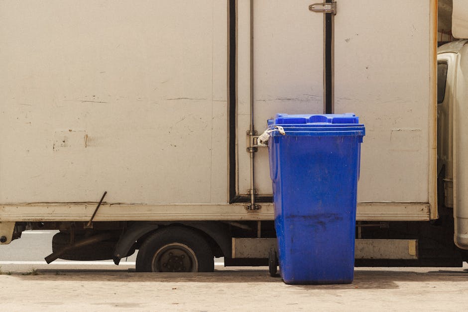 A white delivery van parked on a street with its side door closed, featuring a large blue waste bin positioned on the pavement directly in front of it. The waste bin is made of plastic, with a lid and two small wheels at the base, and appears to be slightly dirty. The van's rear wheel and part of its undercarriage are visible, along with the bottom edge of the vehicle's body. The setting suggests a loading or unloading area associated with home relocation or furniture transport, with the focus on the process of moving or waste disposal related to house removals. The environment is well-lit, with no occupants visible, emphasizing the equipment used during a professional moving service, such as packing, loading, or waste management, aligned with the themes of property transfer and logistical planning, as outlined on hounslowmanandvan.org.uk.