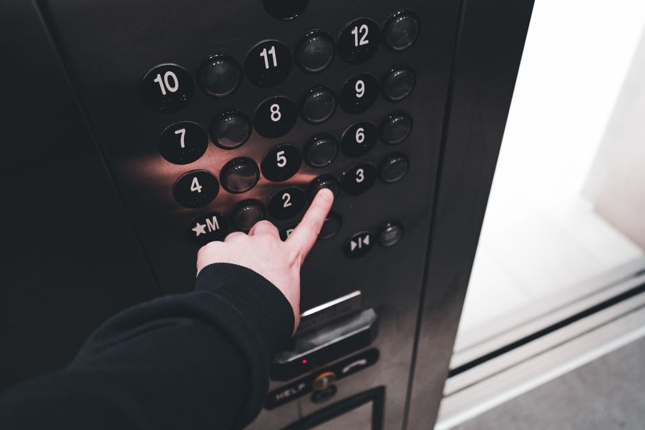 A close-up photograph of a person's hand pressing the number 3 button on an elevator control panel situated inside a building. The panel features multiple black, circular buttons with white numbering from 1 to 12, along with additional symbols such as a star and letter M. The person is wearing a black long-sleeved garment, and the perspective shows the hand reaching out toward the panel, which is mounted on a dark-colored wall. The elevator door is partially visible on the right side of the image. This scene captures the process of selecting a floor during a home relocation or moving process, emphasizing the importance of accessibility and navigation within building environments, an aspect often addressed by removals services like those provided by Hounslow Man and Van.