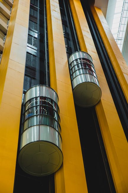 Two glass-fronted elevator cabins with cylindrical shape and metallic accents are installed inside a modern building with yellow walls and large glass windows. The elevators are positioned adjacent to each other, with the doors visible on the side facing the viewer, and are supported by metal tracks embedded in the wall. The surrounding environment is well-lit with natural light coming through the window panels above, highlighting the sleek design of the elevators and the polished surface of the glass. The image captures the internal shaft area dedicated to residential or commercial building logistics, emphasizing the importance of functional and accessible lift systems during home relocation or furniture transport processes, which [COMPANY_NAME], such as Hounslow Man and Van, can assist with for effective removal services.