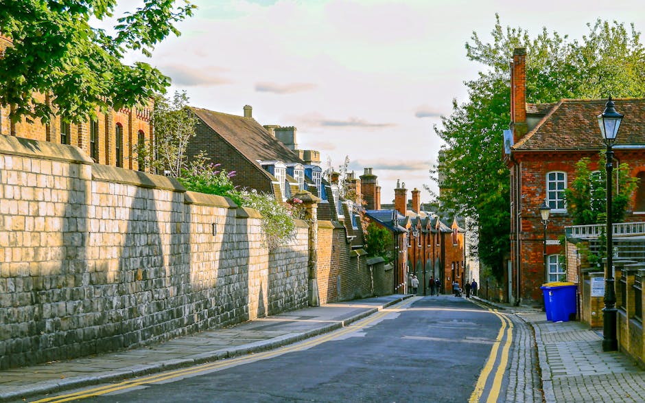 A narrow residential street in Hounslow with a sloped asphalt road featuring double yellow lines along the edge. To the left, there is a high stone wall made of large, light-colored bricks, partially shaded by trees with green foliage. Behind the wall, traditional brick houses with pitched roofs and chimneys are visible. On the right side, a red brick building with white-framed windows and a small balcony is adjacent to a tall, black street lamp and a blue recycling bin placed on the sidewalk. Further down the street, a group of people dressed in casual clothing are seen walking or standing near the storefronts. The scene is illuminated by natural daylight, with a partly cloudy sky overhead, highlighting the historical architecture and the urban environment typical of a house relocation or moving process environment as provided by Hounslow Man and Van.