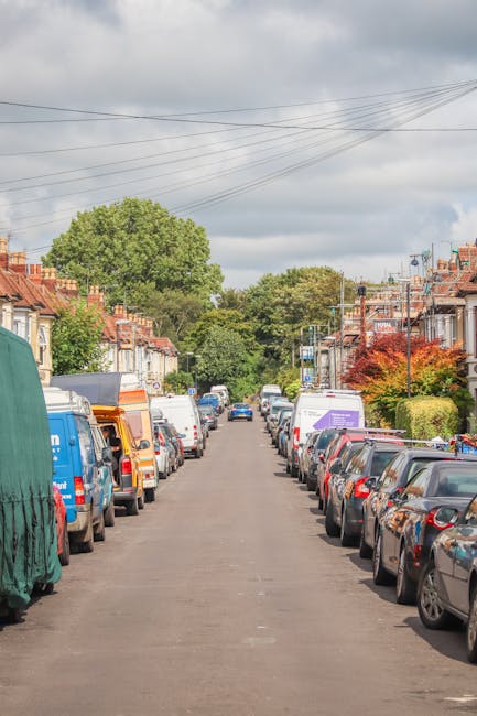 A narrow residential street in Hounslow lined with parked cars on both sides, including sedans, hatchbacks, and small vans, with some vehicles partially covered by protective tarps or blankets. The pavement is bordered by rows of terraced houses with small front gardens, some featuring colorful foliage or bushes. Overhead, multiple power lines stretch across the street beneath a partly cloudy sky. In the distance, a Hounslow Man and Van vehicle is preparing to load furniture and packed boxes into the van using lifting equipment, as part of a home relocation or moving process. The scene illustrates the logistical challenges faced during house removals in tight, urban neighborhoods, highlighting the importance of careful planning and specialized transport services such as those provided by Hounslow Man and Van, to facilitate furniture transport and packing and moving tasks in constrained street environments.