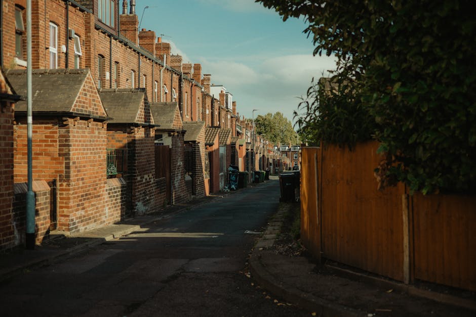 A narrow residential street in Hounslow with a sloped asphalt road featuring double yellow lines along the edge. To the left, there is a high stone wall made of large, light-colored bricks, partially shaded by trees with green foliage. Behind the wall, traditional brick houses with pitched roofs and chimneys are visible. On the right side, a red brick building with white-framed windows and a small balcony is adjacent to a tall, black street lamp and a blue recycling bin placed on the sidewalk. Further down the street, a group of people dressed in casual clothing are seen walking or standing near the storefronts. The scene is illuminated by natural daylight, with a partly cloudy sky overhead, highlighting the historical architecture and the urban environment typical of a house relocation or moving process environment as provided by Hounslow Man and Van.
