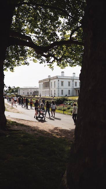 A group of people walking along a wide paved path through Osterley Park, with some pushing prams and others carrying bags, in front of a large, white neoclassical building with tall windows and a flat roof. The scene is shaded by a large tree with leafy branches that extend overhead, casting dappled sunlight across the pathway. The park features well-maintained lawns and flowerbeds on either side of the path, with a low metal fence enclosing the area. This outdoor setting illustrates a typical day of visitors enjoying the park’s open space. The image is relevant to house removals and relocation services, such as those provided by Hounslow Man and Van, highlighting outdoor environments that clients may need to access or prepare for during home relocation or furniture transport processes, especially when planning routes and timing to Osterley Park during a house move.