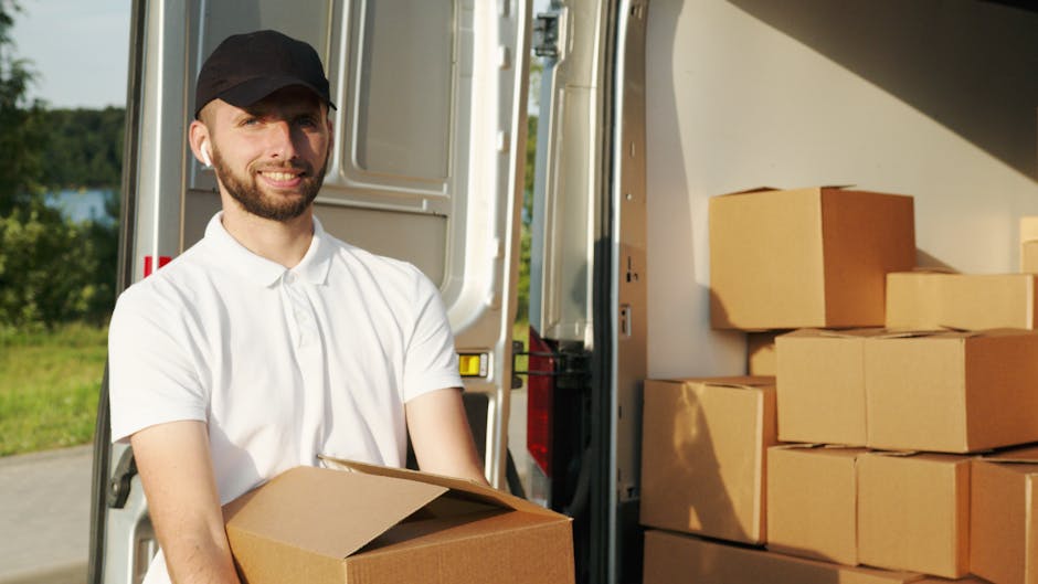 A man with a beard, wearing a black cap and a white polo shirt, is pictured outdoors next to a moving van during the loading process. He is holding a cardboard box and smiling at the camera. Inside the van, there are several other cardboard boxes stacked on the floor and against the side walls, some wrapped in plastic or packing materials, indicating a home relocation. The background includes a green landscape with trees and a body of water, under natural lighting. The scene reflects the typical packing and furniture transport involved in same day removals, with visible packaging materials and equipment such as the moving van's interior and a trolley nearby, illustrating efficient logistics managed by Hounslow Man and Van for local removals.