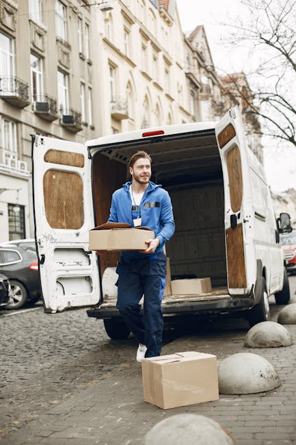 A male mover with a beard, wearing a blue jacket and dark work trousers, is standing at the back of a white moving van parked on a cobblestone street in an urban residential area. He is holding a medium-sized cardboard box and appears to be in the process of loading or unloading it. The van's rear doors are open, revealing an empty wooden interior suitable for transporting household furniture and cartons. On the ground beside him, there is one additional cardboard box and two large rounded concrete bollards. The background features multi-storey buildings with classic architectural details, including windows with black railings and decorative facades. The scene is set during daytime with natural light, illustrating the typical logistics involved in home relocation, furniture transport, and packing and moving operations, as handled by companies like Hounslow Man and Van.