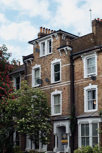 Photograph of a traditional multi-storey brick residential building with white window frames and decorative trim, situated on a street with greenery including a tree with branches and leaves visible in the foreground. Satellite dishes are attached to the exterior walls, and a sign for Hounslow Man and Van is partially visible at the bottom, indicating the location. The building features chimneys on the roof and is illuminated by natural daylight, with a partly cloudy sky overhead. This image relates to house removals and furniture transport, highlighting the exterior environment associated with residential moves in Hounslow, suitable for a guide on estate removals for small flats.