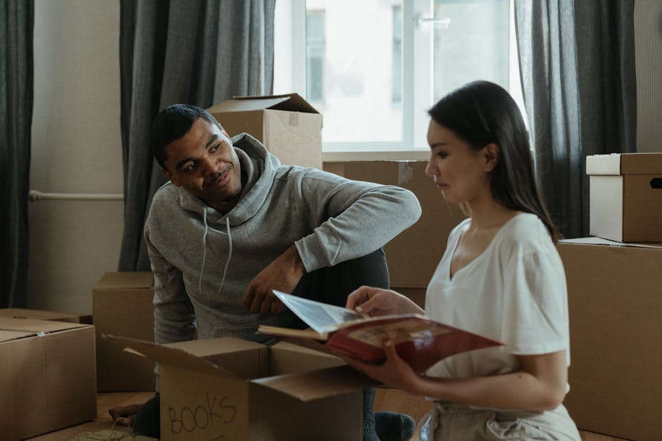 A man and a woman inside a room during a home relocation process, surrounded by packed cardboard boxes of various sizes, some labeled with contents such as 'books.' The man, dressed in a gray hoodie, is leaning forward with a slight smile, while the woman, in a white top, is seated and reading a document or instruction sheet. Natural light filters through a large window behind them, with dark curtains partially drawn, illuminating the space that reflects typical packing and moving activities. The scene depicts the packing and organizing phase of furniture transport and house removals, with visible packaging materials like cardboard and plastic wrap, and furniture such as a cardboard box and a possible small piece of furniture nearby. This image captures the preparatory steps involved in professional removals, echoing the logistics and packing challenges often encountered during house moves in the Hounslow area, as referenced in the page about weekend booking delays and common problems for Hounslow removals.
