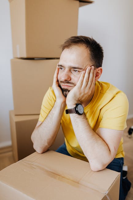 A man in a yellow t-shirt with a wristwatch is sitting at a table indoors, resting his head in his hands with a bored or frustrated expression on his face. Behind him, there are several stacked cardboard boxes, some open and others sealed, indicating the process of home relocation or packing. The room has neutral-colored walls and natural lighting, with a focus on the man's forlorn mood amid the presence of packing materials and boxes, reflecting common problems during house removals. This scene illustrates the emotional and logistical challenges associated with furniture transport and moving processes managed by companies like Hounslow Man and Van, as they handle packing, loading, and transportation tasks during home moves.